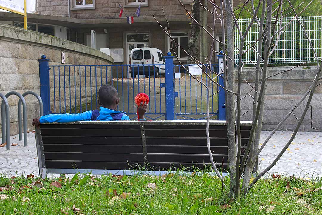 Portrait de dos devant l'entrée d'une école. Enfant sur un banc, main avec Pompon rouge. Zone Artistique de Pomponnerie, Quartier Frébault-Polygone Lorient, Art participatif, Viviane Rabaud