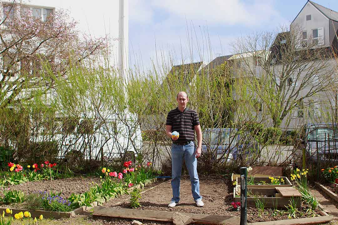 Portrait. Adulte debout dans jardin potager, fleurs, main avec Pompon bleu clair. Zone Artistique de Pomponnerie, Quartier Frébault-Polygone Lorient, Art participatif, Viviane Rabaud