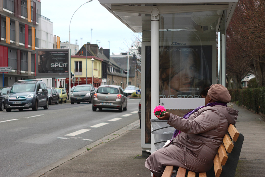 Portrait. Femme sur un banc dans abri-bus, main avec Pompon rose et vert. Zone Artistique de Pomponnerie, Quartier Frébault-Polygone Lorient, Art participatif, Viviane Rabaud
